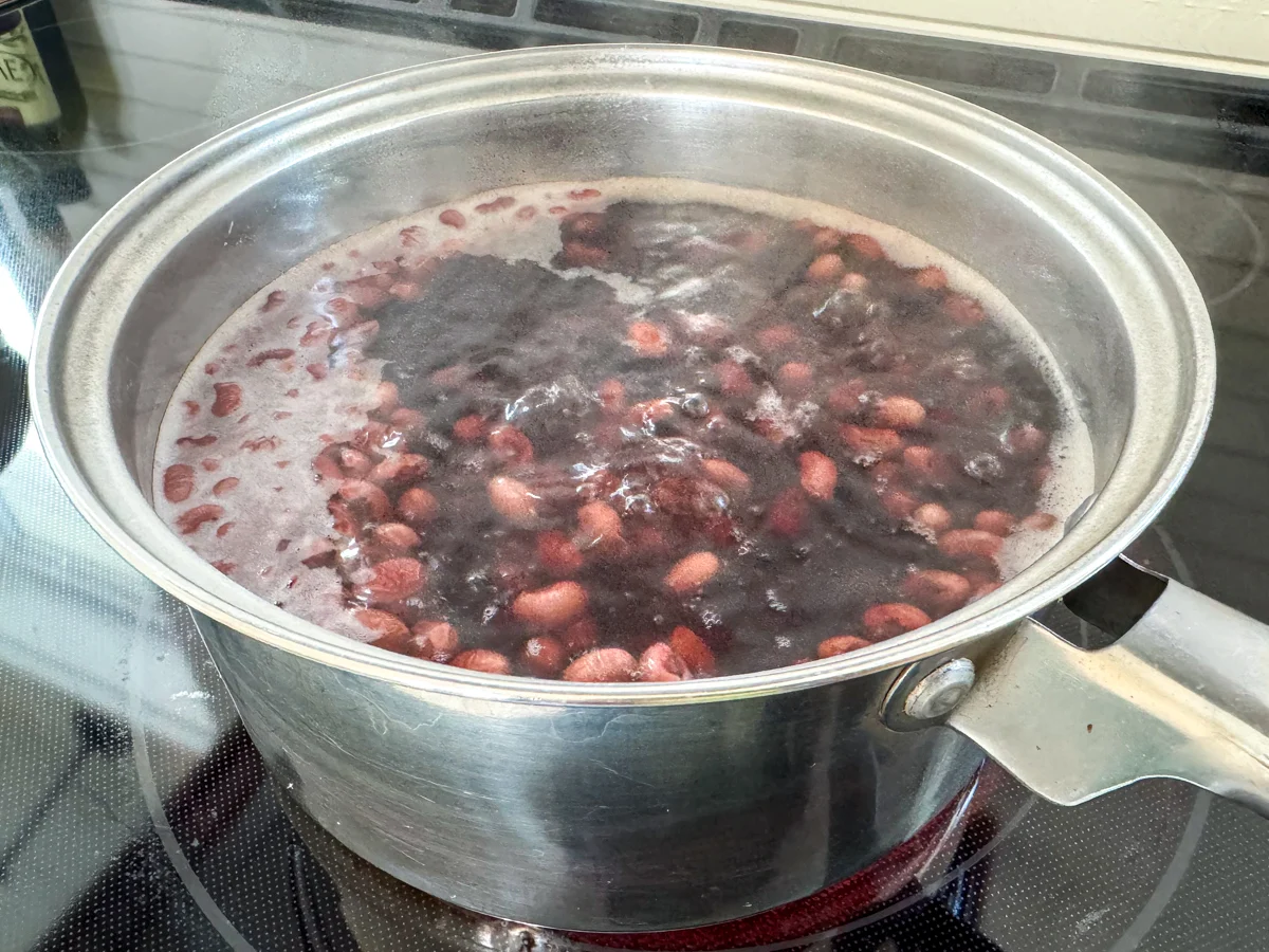 A Dutch oven on a stovetop with dried beans gently simmering in clear broth, a wooden spoon resting on the rim