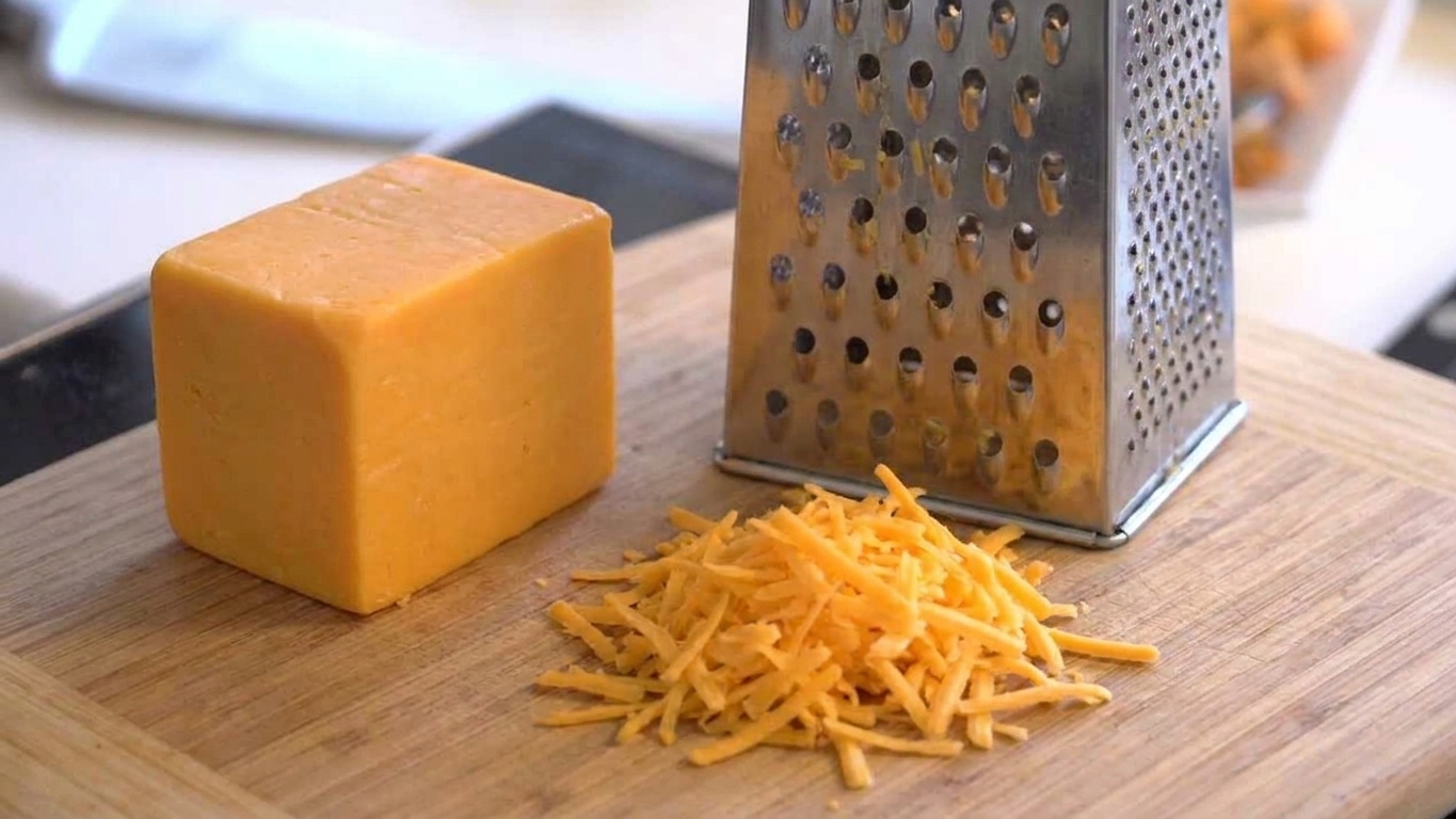 A block of cheddar next to a box grater with a small pile of freshly grated cheese on a cutting board