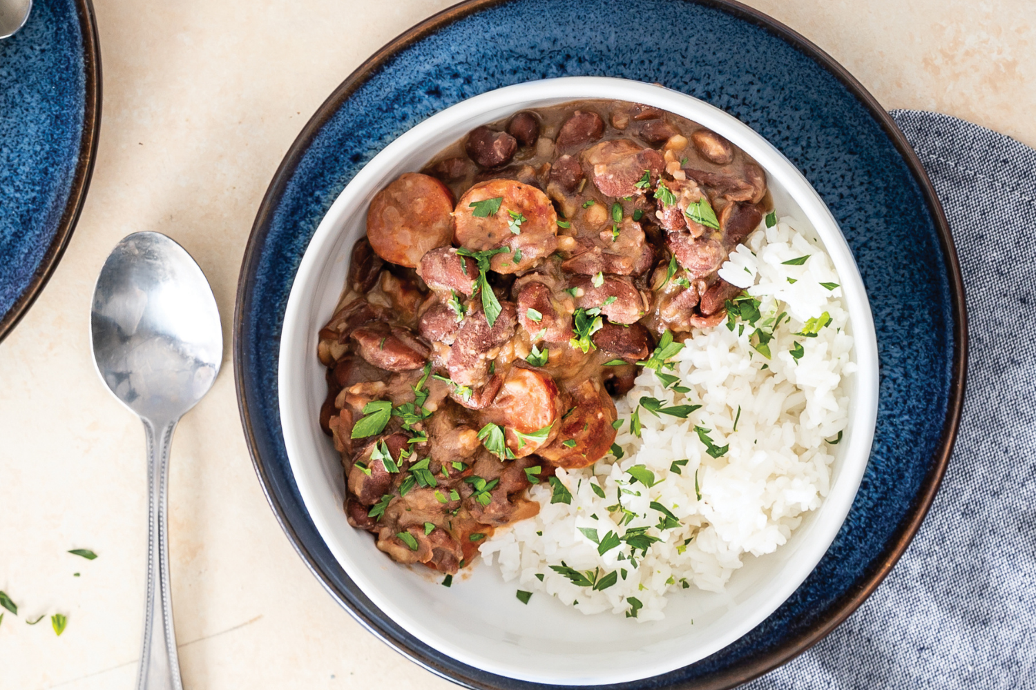 A bowl of Louisiana-style red beans and creamy bean gravy spooned over white rice, topped with sliced smoked sausage on a wooden table with a spoon nearby