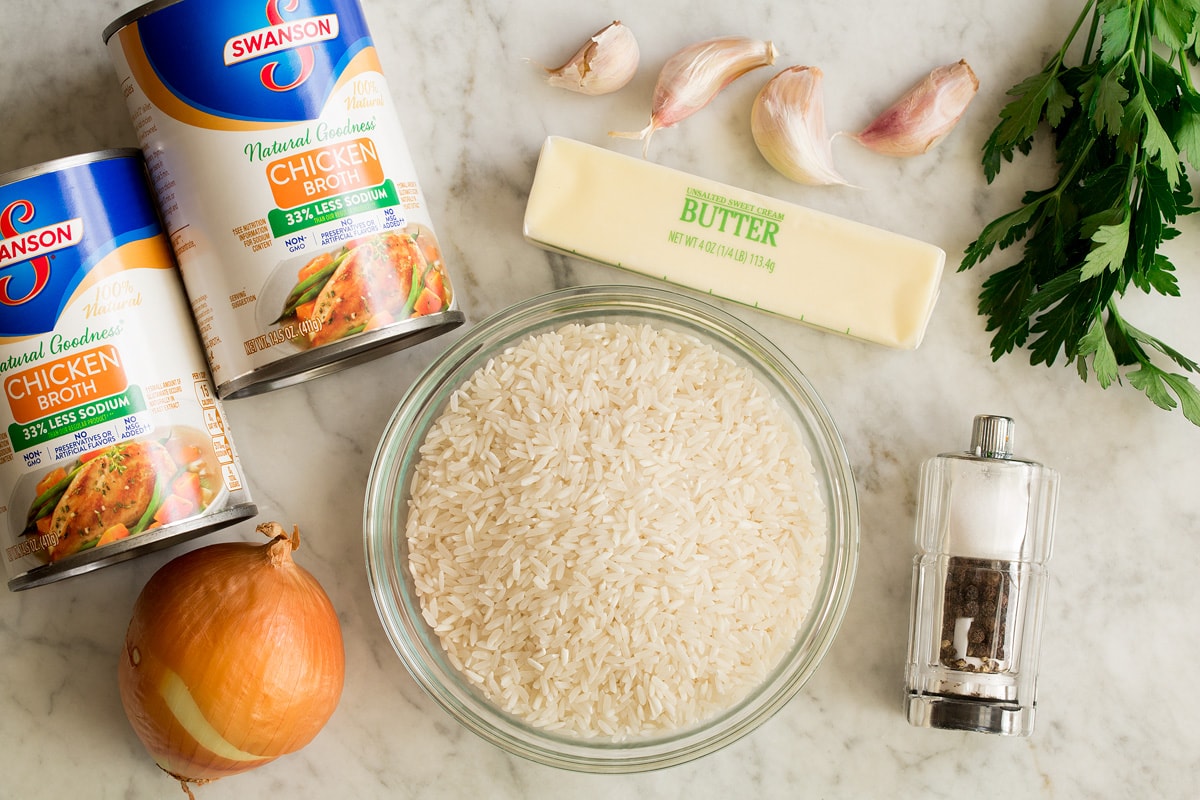 A bowl of uncooked long-grain white rice on a counter next to garlic cloves and butter.
