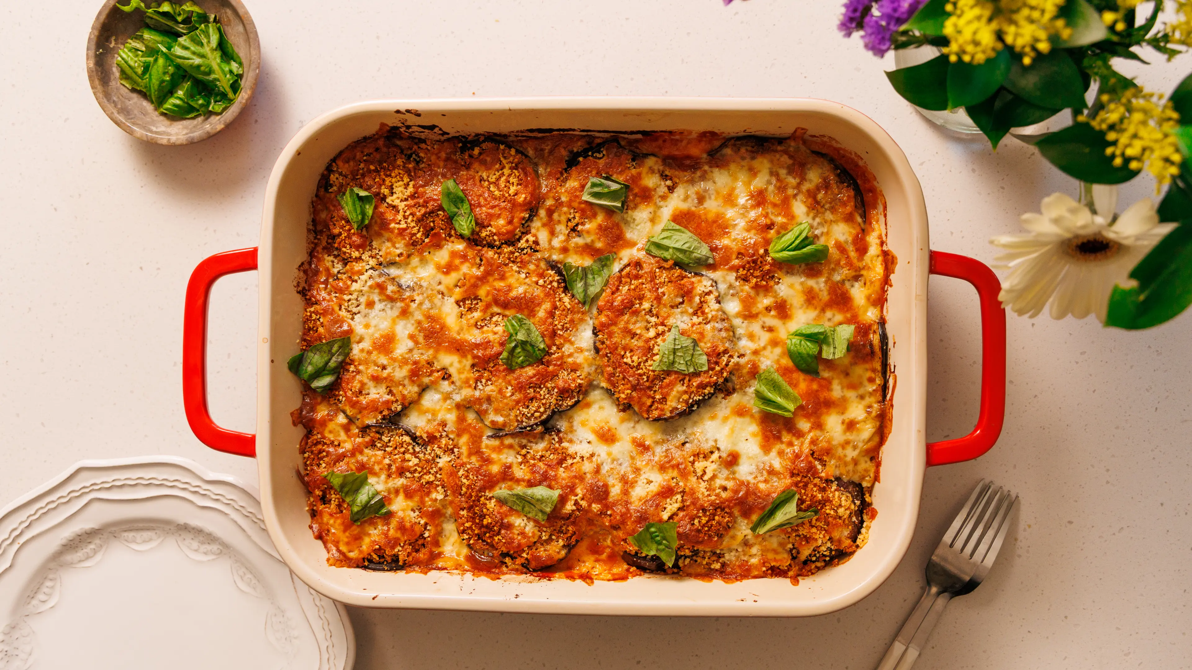 A bubbling pan of baked eggplant Parmesan with browned mozzarella, scattered basil, and crisp breaded eggplant edges, photographed on a kitchen counter