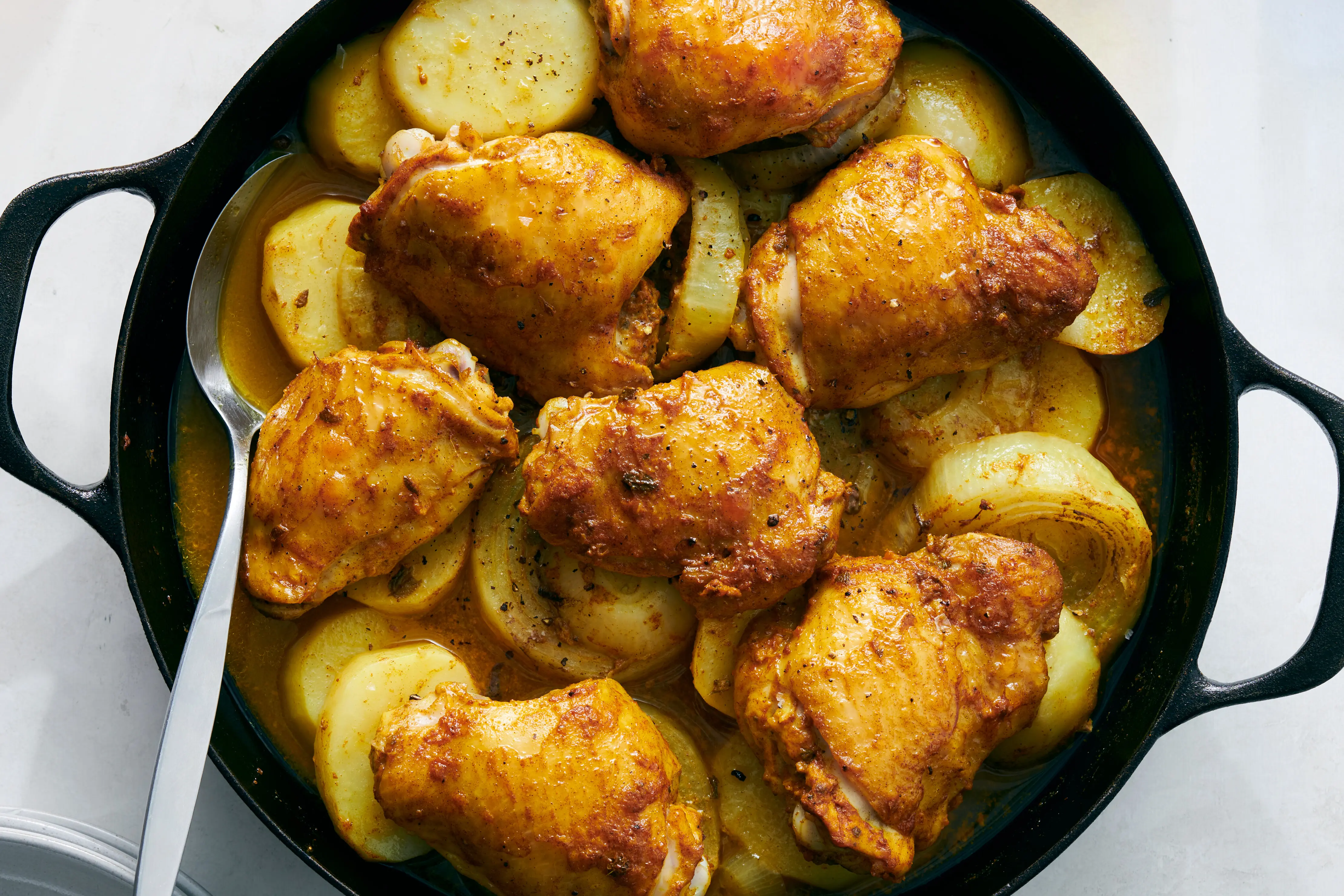 A cast iron skillet on a stove with browned chicken pieces simmering in a tomato sauce, with a wooden spoon resting nearby