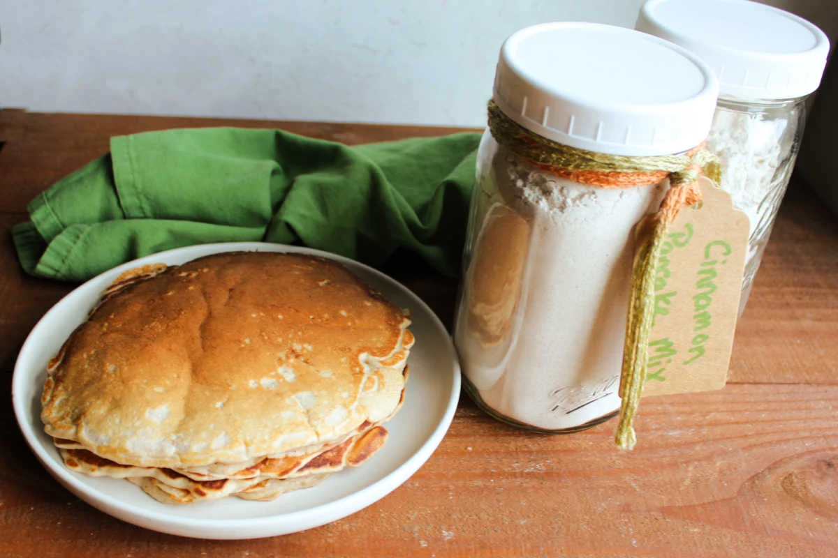 A clear glass jar filled with homemade pancake mix on a kitchen counter, with a whisk and measuring spoons nearby