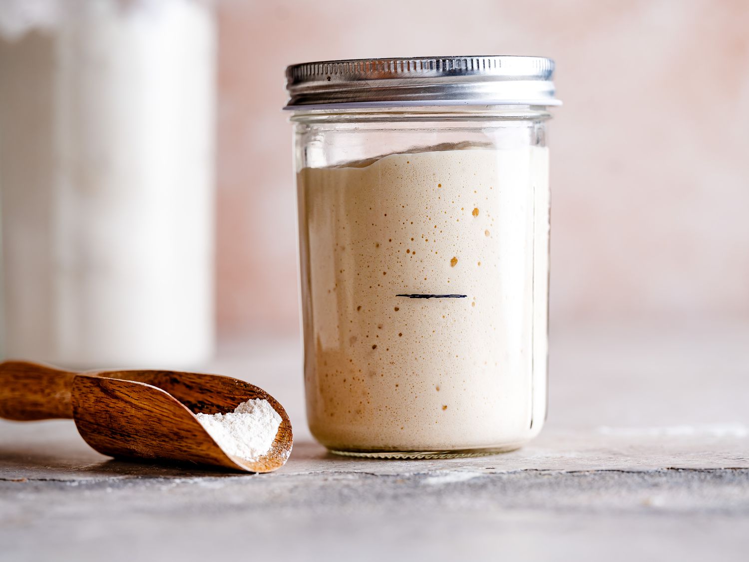 A clear glass jar of active sourdough starter with bubbles and a visible rise line, sitting on a clean kitchen counter near a small bowl of flour