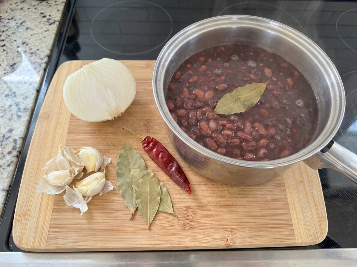 A cutting board with a halved onion, smashed garlic cloves, bay leaves, and a dried chile arranged next to a pot