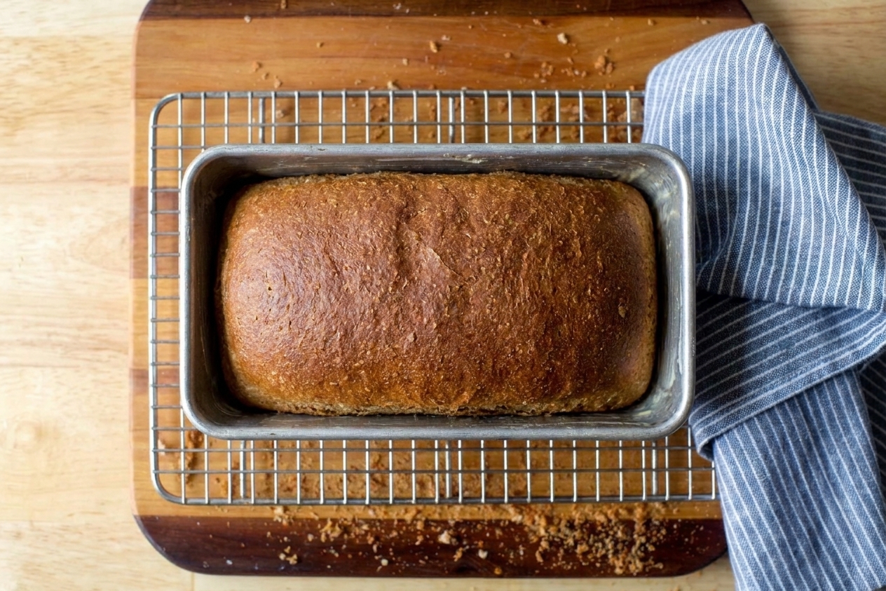 A deep golden wheat bread loaf sitting in a metal pan on a cooling rack
