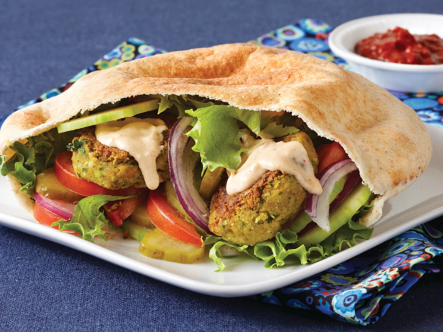 A falafel pita sandwich filled with chopped tomatoes, cucumbers, lettuce, and a drizzle of tahini sauce on parchment paper