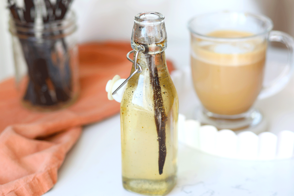 A glass jar of homemade vanilla syrup with a small spoon on a wooden kitchen counter in soft window light