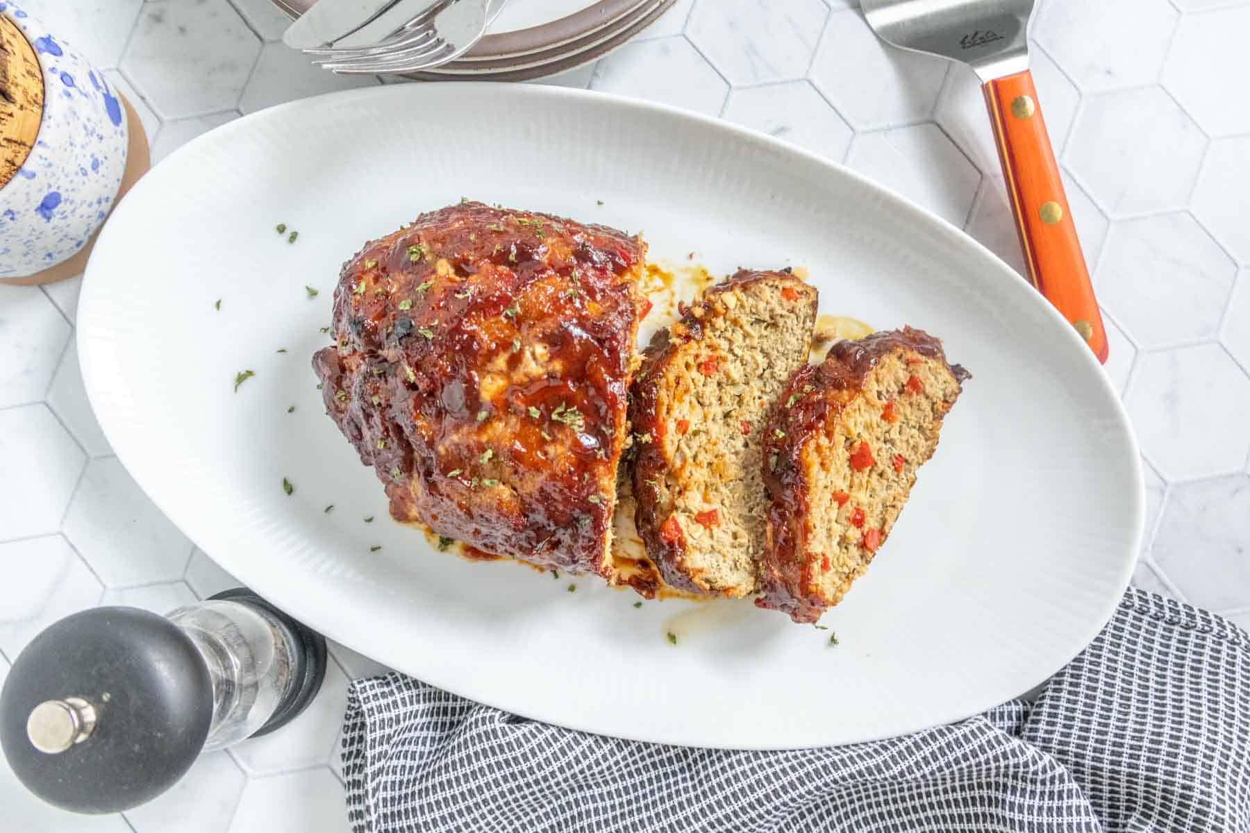 A glossy, ketchup-glazed meatloaf sliced into thick pieces on a warm platter with juices pooling slightly, dinner table setting