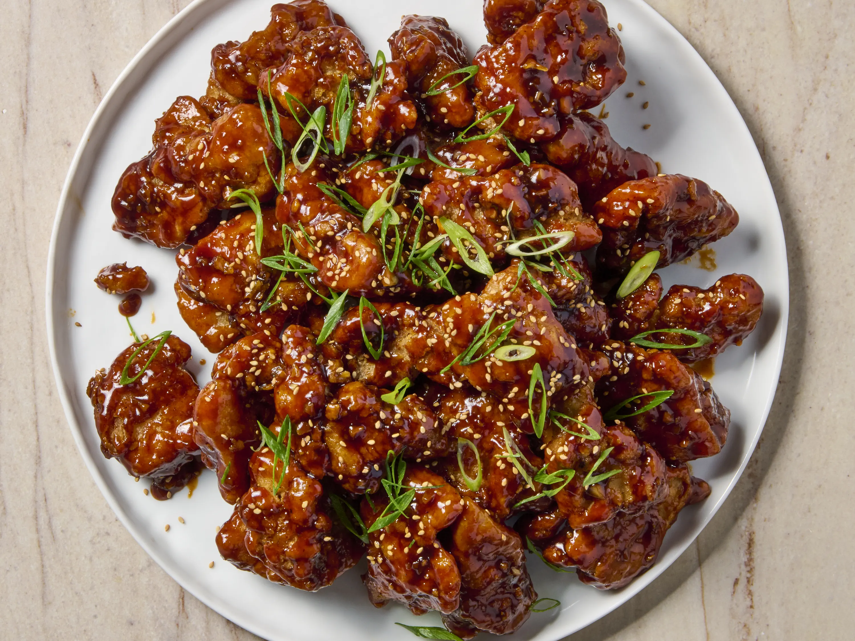 A glossy platter of Korean BBQ chicken with caramelized edges, sliced scallions, toasted sesame seeds, and a small bowl of extra sauce on the side