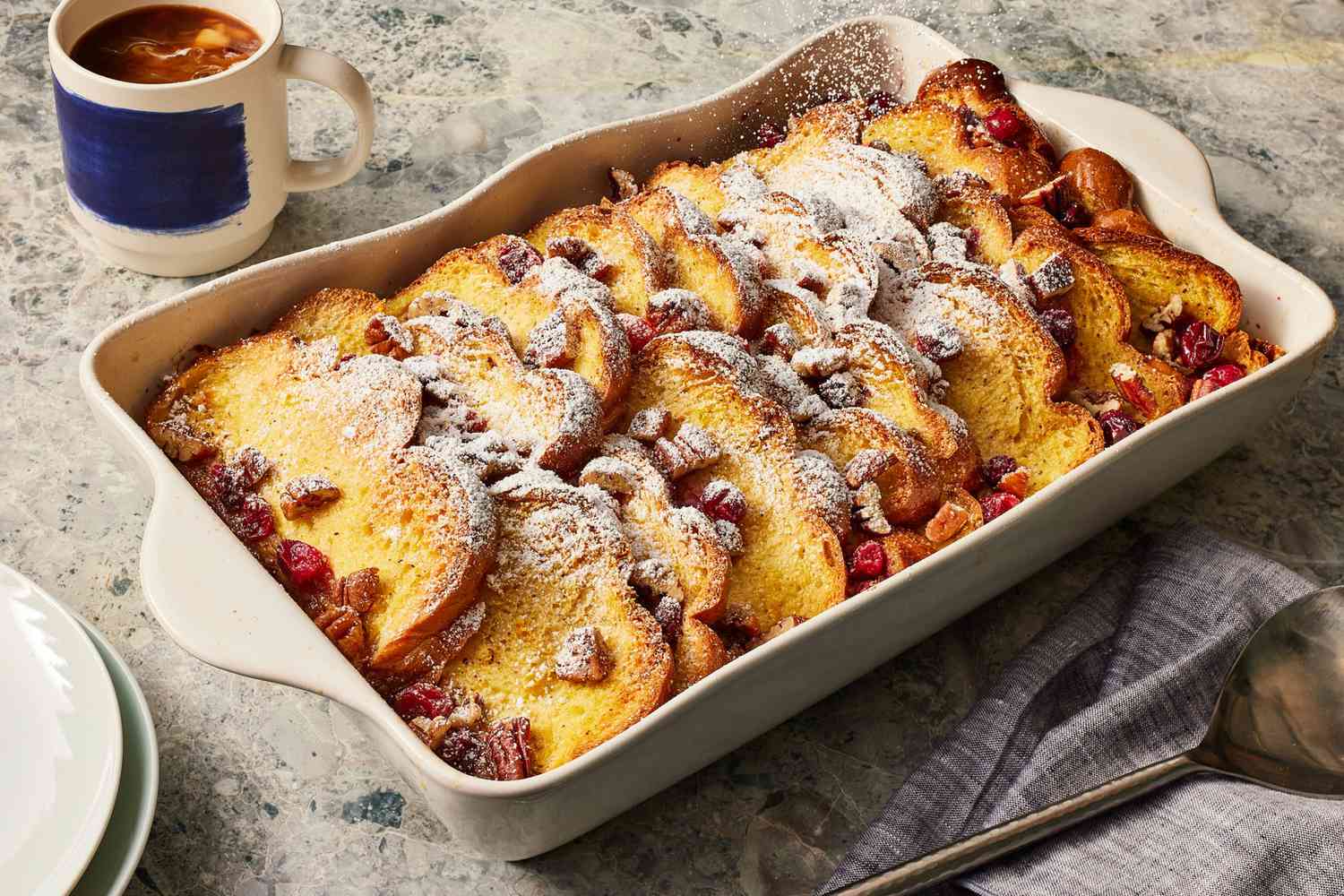 A golden-brown baked French toast casserole in a ceramic baking dish on a wooden table, with crisp edges and powdered sugar lightly dusted on top