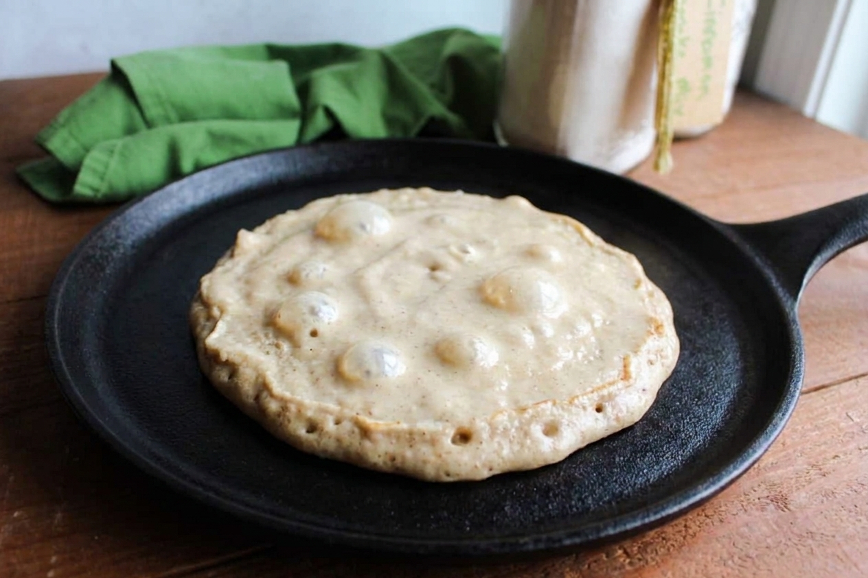 A pancake cooking on a griddle with bubbles forming on the surface and edges beginning to set