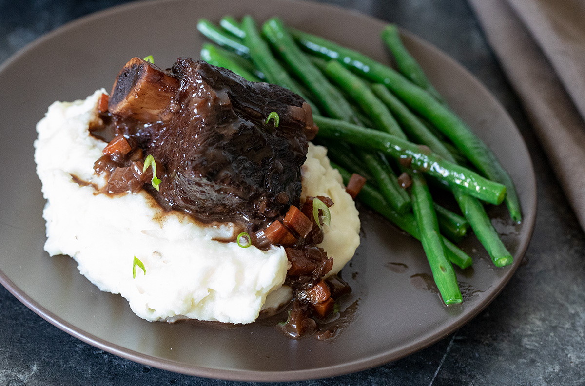 A platter of braised beef short ribs coated in glossy sauce, with carrots and herbs scattered around, set on a rustic dinner table