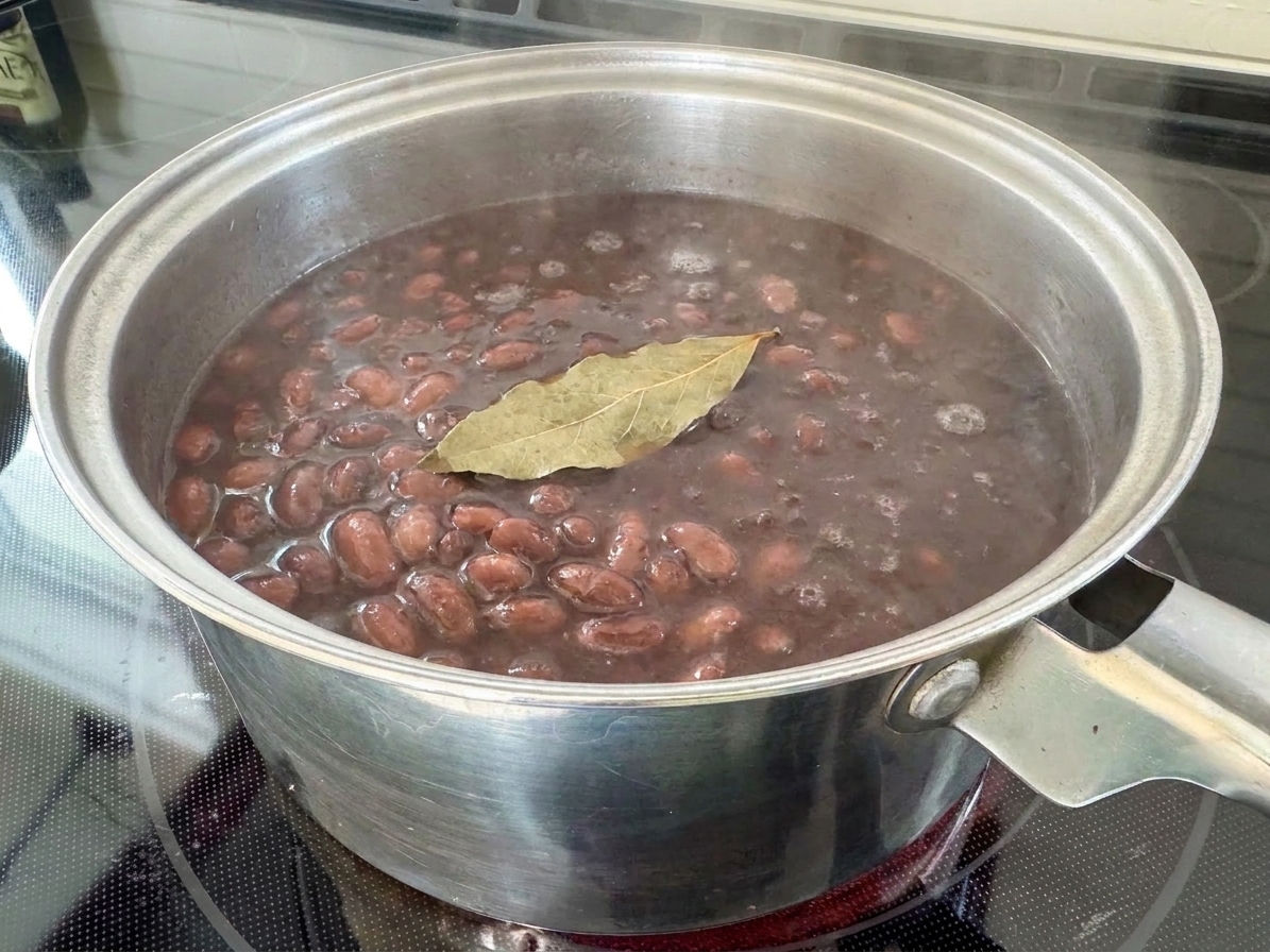 A pot of cooked black beans in their dark broth with a bay leaf floating on top
