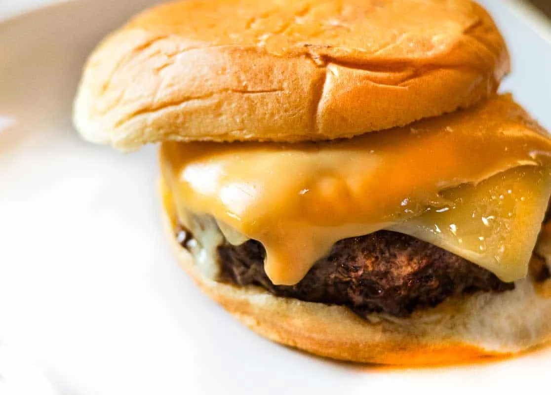 A rustic homestyle burger on a toasted bun with melted cheddar, pickles, onion, and a smear of sauce on a wooden board with a cast iron skillet in the background