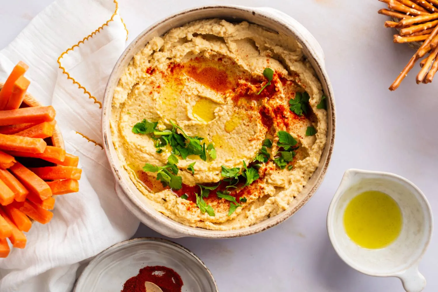 A shallow bowl of extra creamy hummus swirled with a spoon, topped with olive oil, paprika, and chopped parsley on a neutral linen table setting
