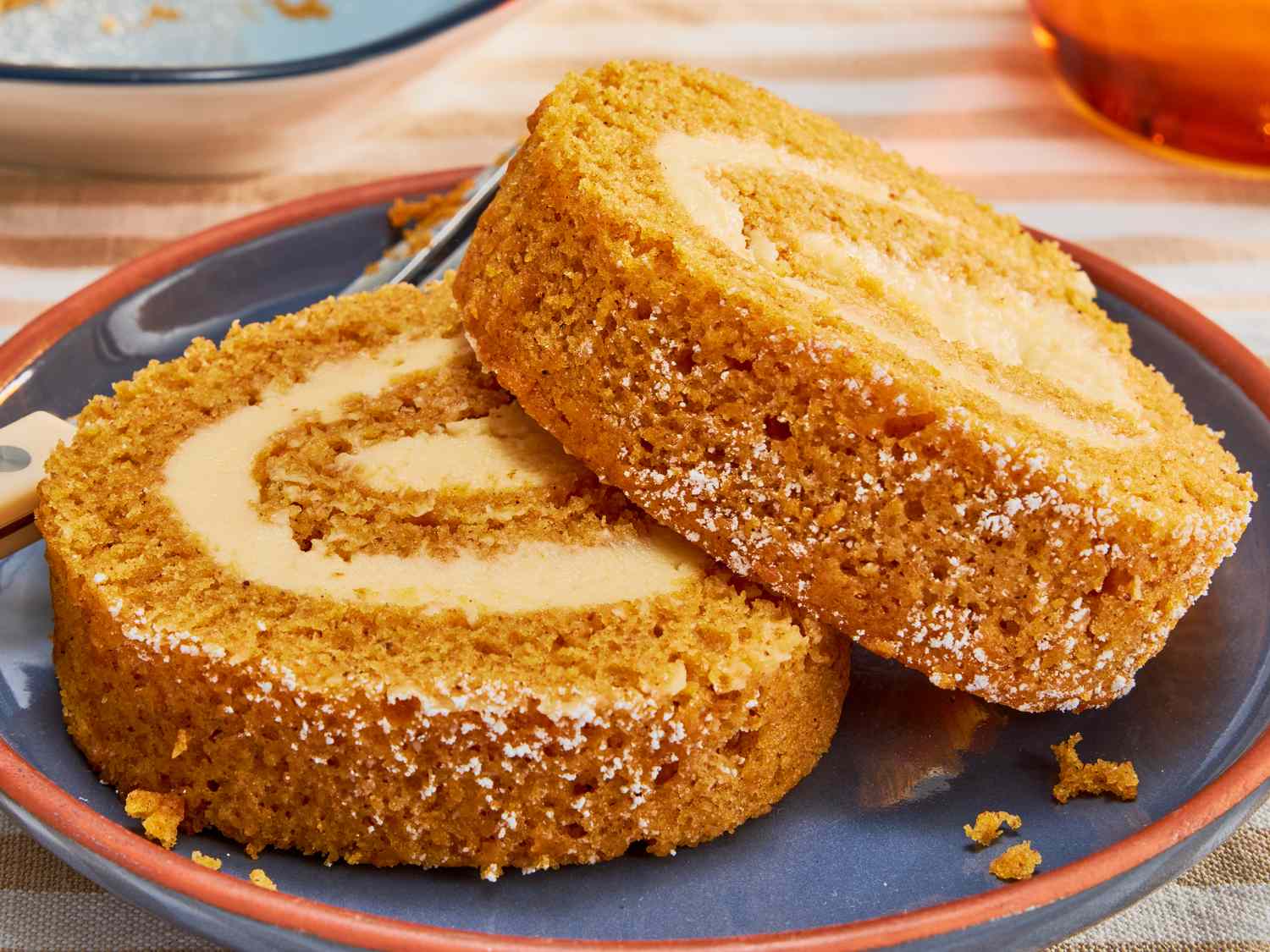 A sliced pumpkin roll on a ceramic plate, showing a tight cream cheese swirl and a light dusting of powdered sugar
