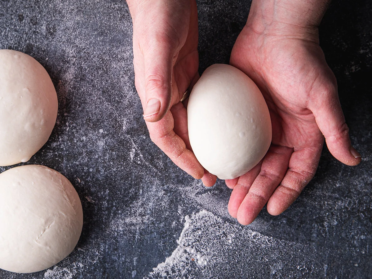 A smooth, round ball of homemade pizza dough resting on a lightly floured wooden countertop with a clean kitchen towel nearby