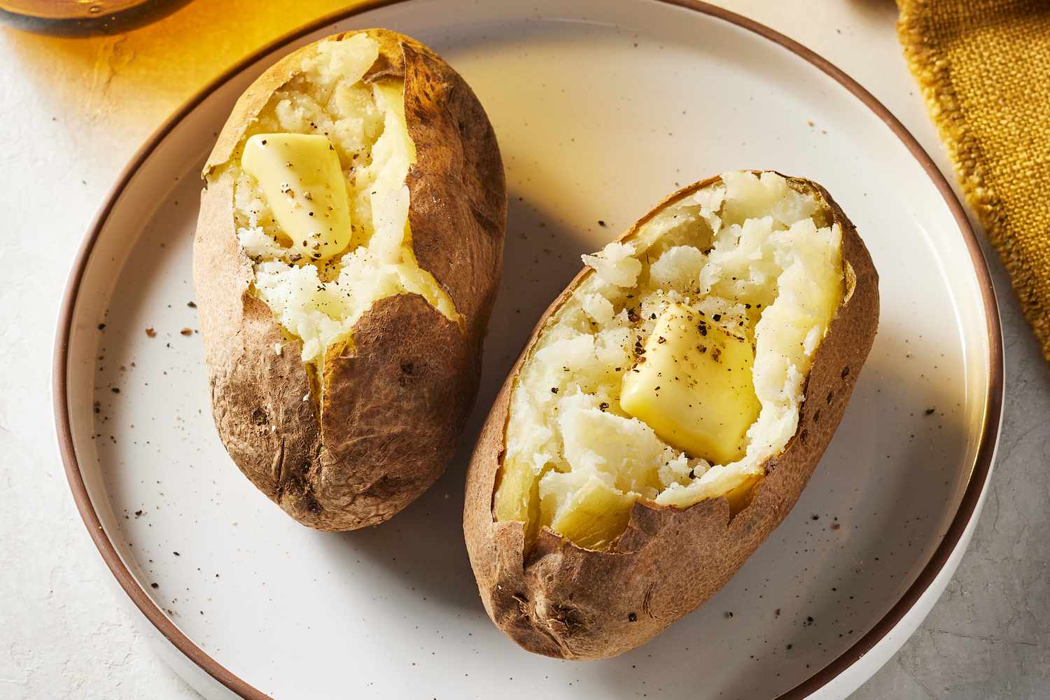 A split open baked russet potato on a white plate with crisp, wrinkly skin and a fluffy steaming interior, topped with a small pat of butter