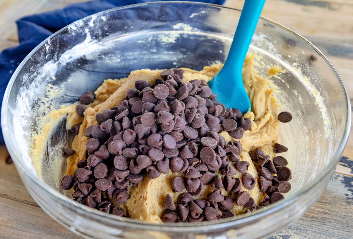 A stainless steel mixing bowl filled with chocolate chip cookie dough on a kitchen counter with a rubber spatula resting on the rim