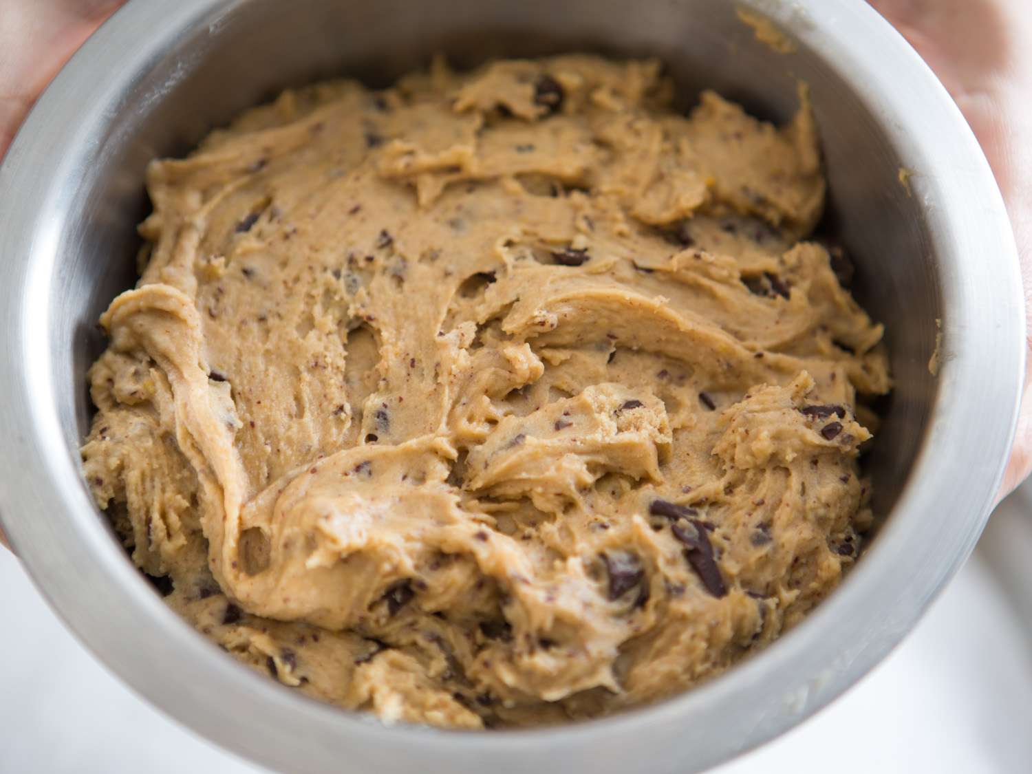 A stainless steel mixing bowl filled with thick chocolate chip cookie dough, with chocolate chips visible throughout on a tidy kitchen counter
