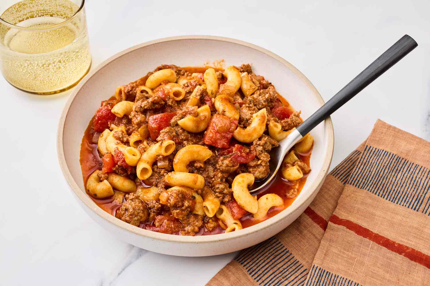 A steaming bowl of American goulash with elbow macaroni, beefy tomato sauce, and a sprinkle of chopped parsley on top, set on a cozy dinner table