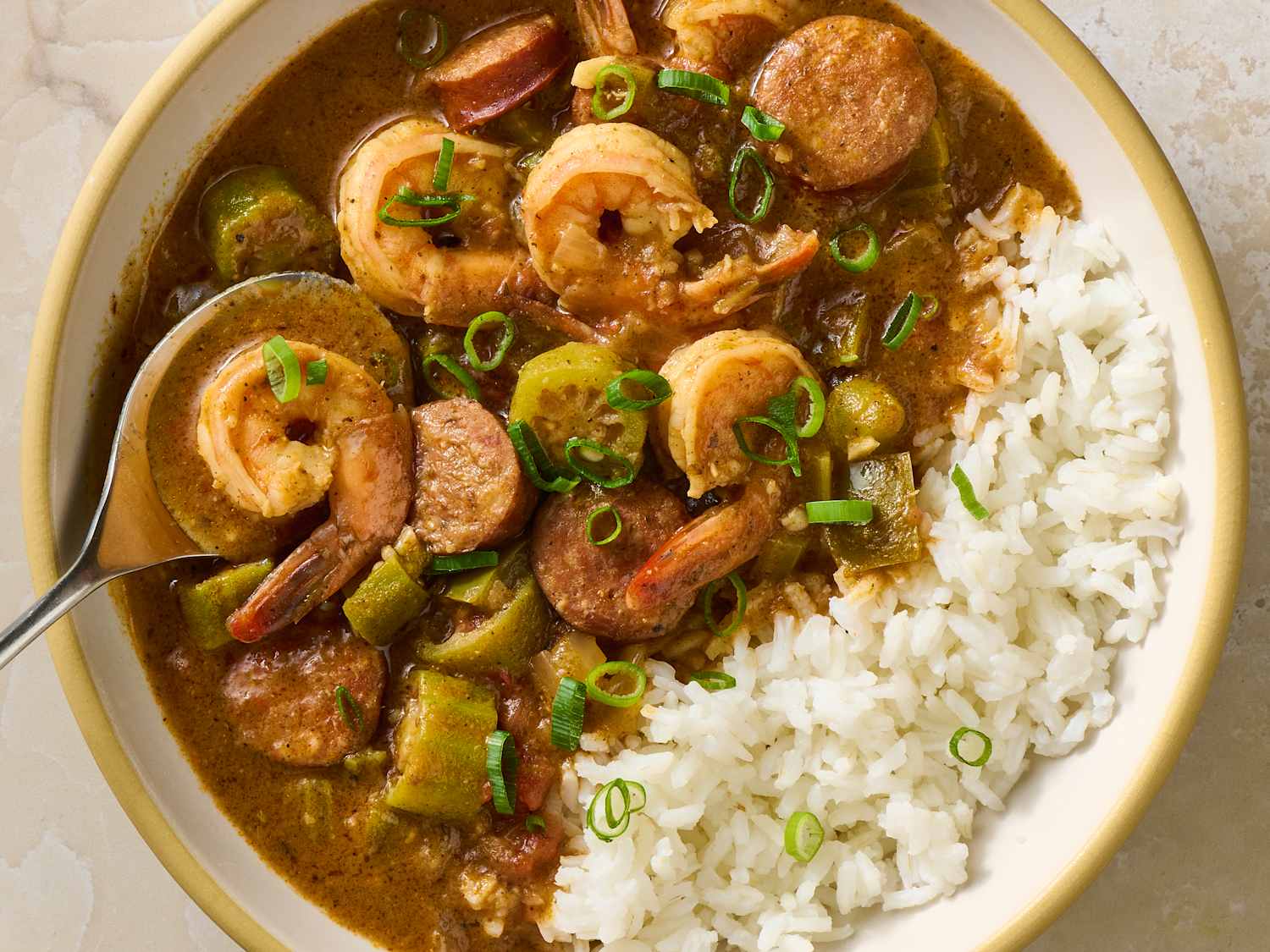 A steaming bowl of shrimp gumbo with white rice in the center, visible pieces of shrimp, okra, and green onions, set on a wooden table with a spoon beside it