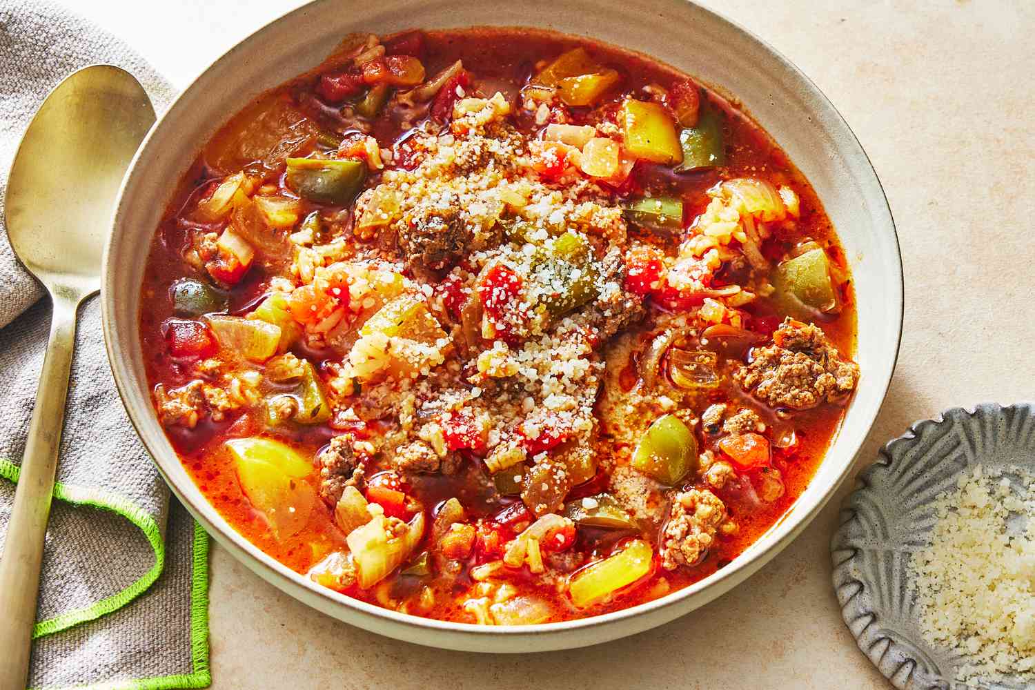 A steaming bowl of stuffed pepper soup with visible pieces of bell pepper, ground beef, rice, and herbs, shot on a wooden table with a spoon nearby