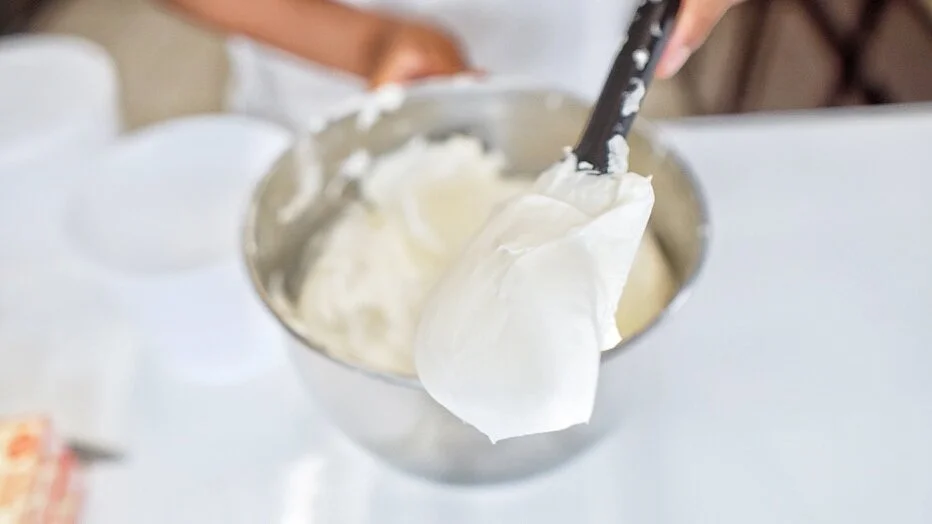 A swirl of smooth, glossy buttercream frosting in a white mixing bowl with a spatula resting on the rim, photographed on a bright kitchen counter