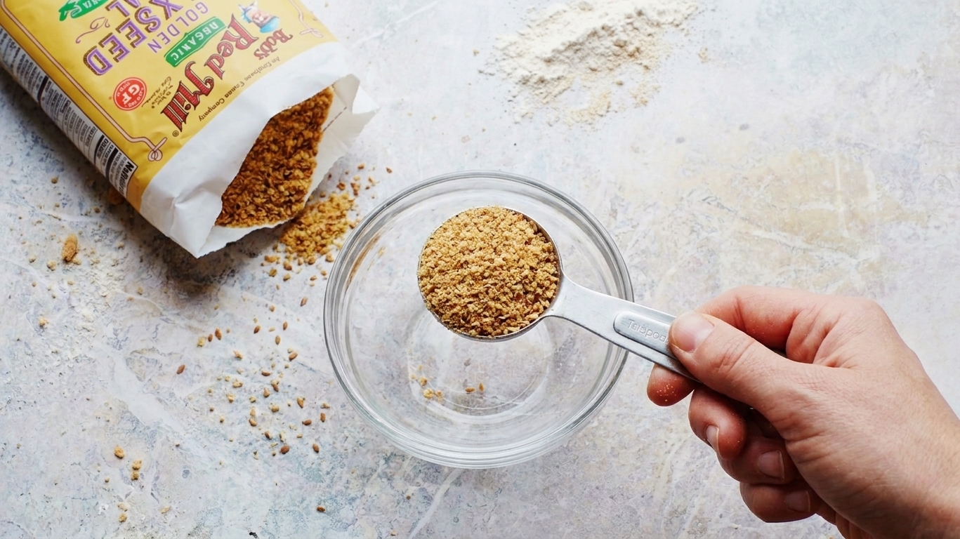 A tablespoon measuring spoon filled with ground flaxseed held over a small bowl on a kitchen counter