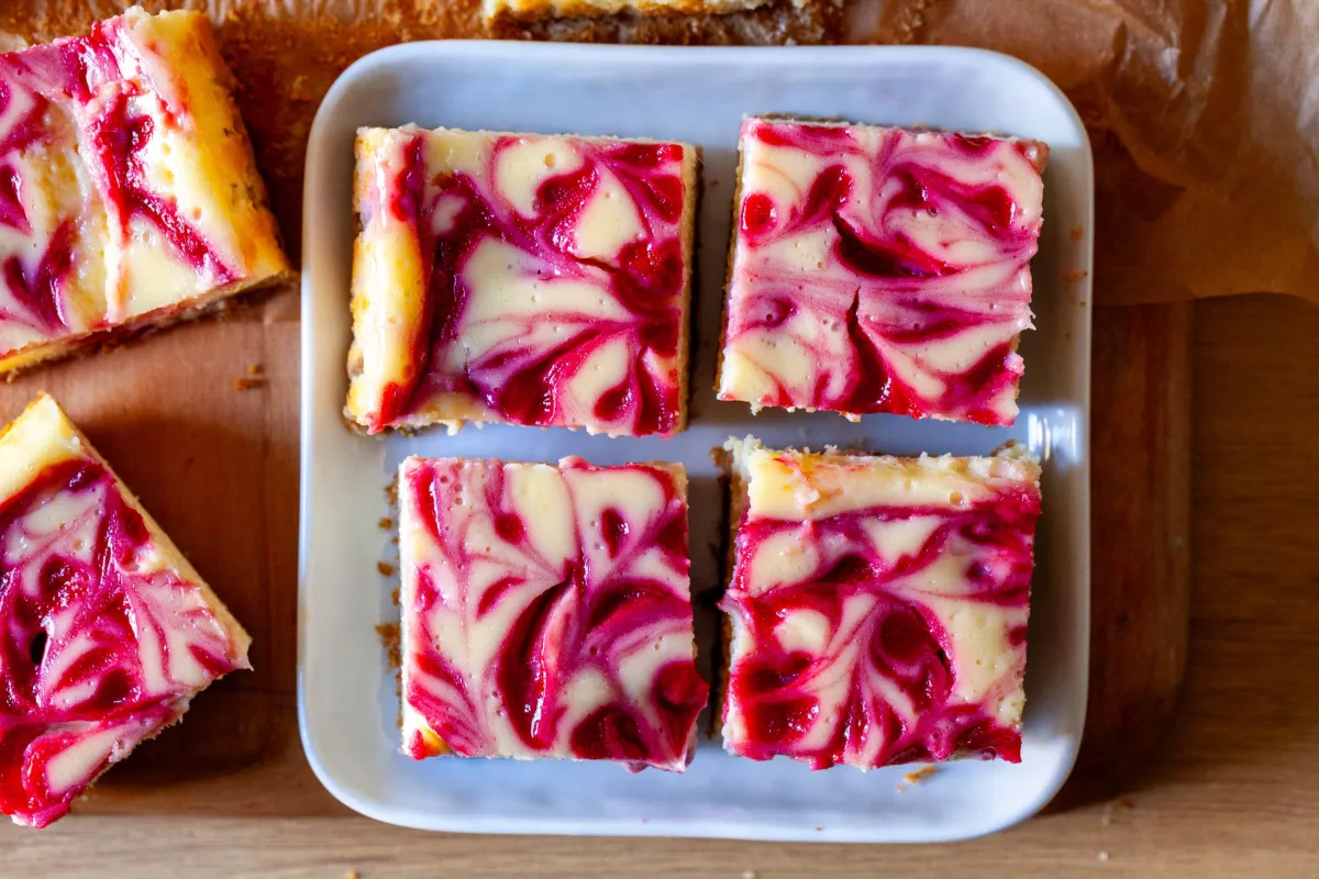 A tall slice of raspberry swirl cheesecake on a small dessert plate, showing creamy filling with vivid raspberry ribbons and a graham cracker crust