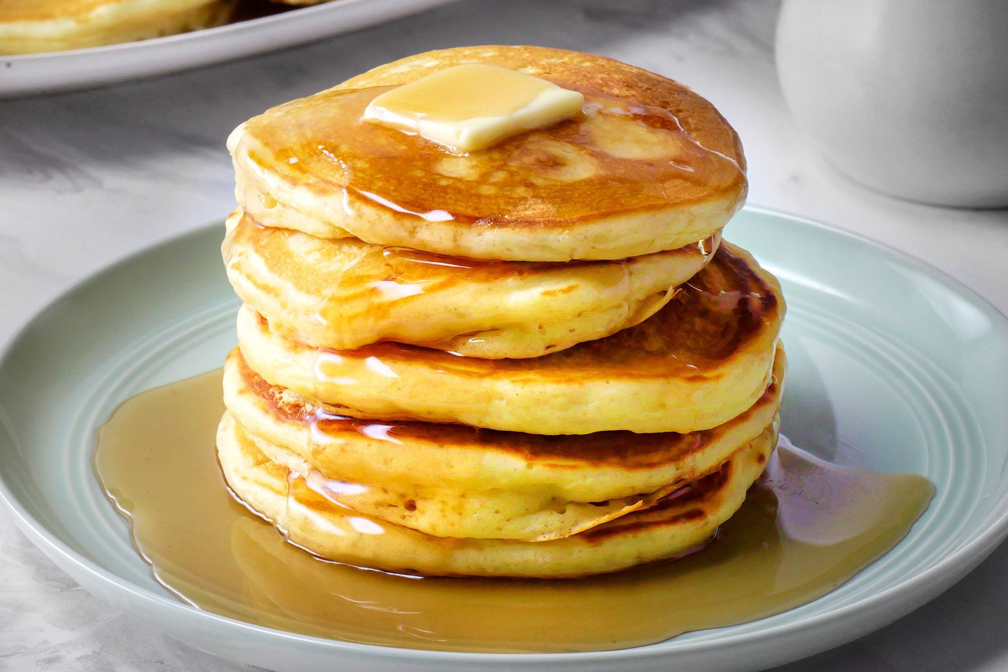 A tall stack of fluffy buttermilk pancakes on a white plate with a pat of butter melting on top and maple syrup drizzled down the sides on a sunlit kitchen table