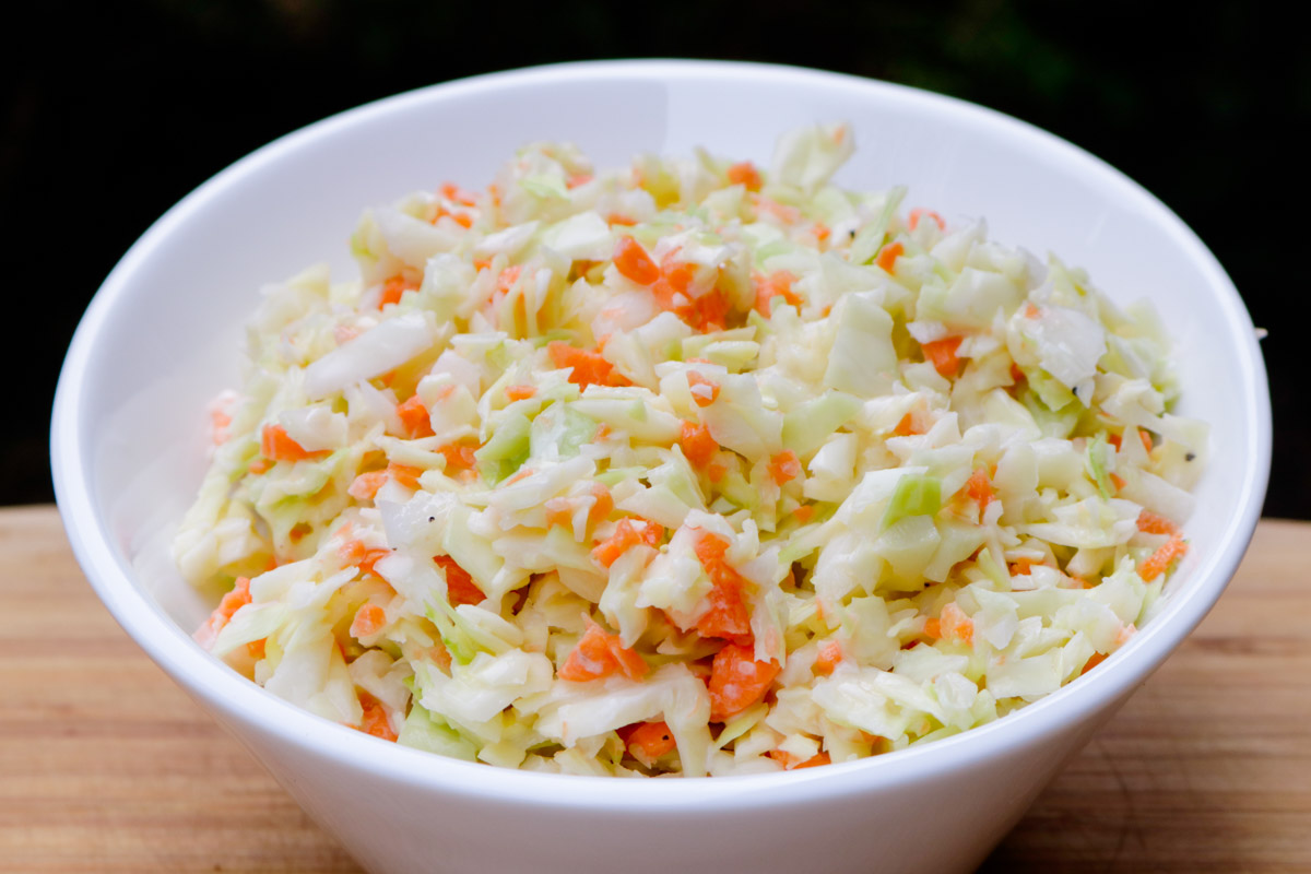 A white bowl filled with creamy finely chopped coleslaw on a kitchen counter, with a serving spoon nearby