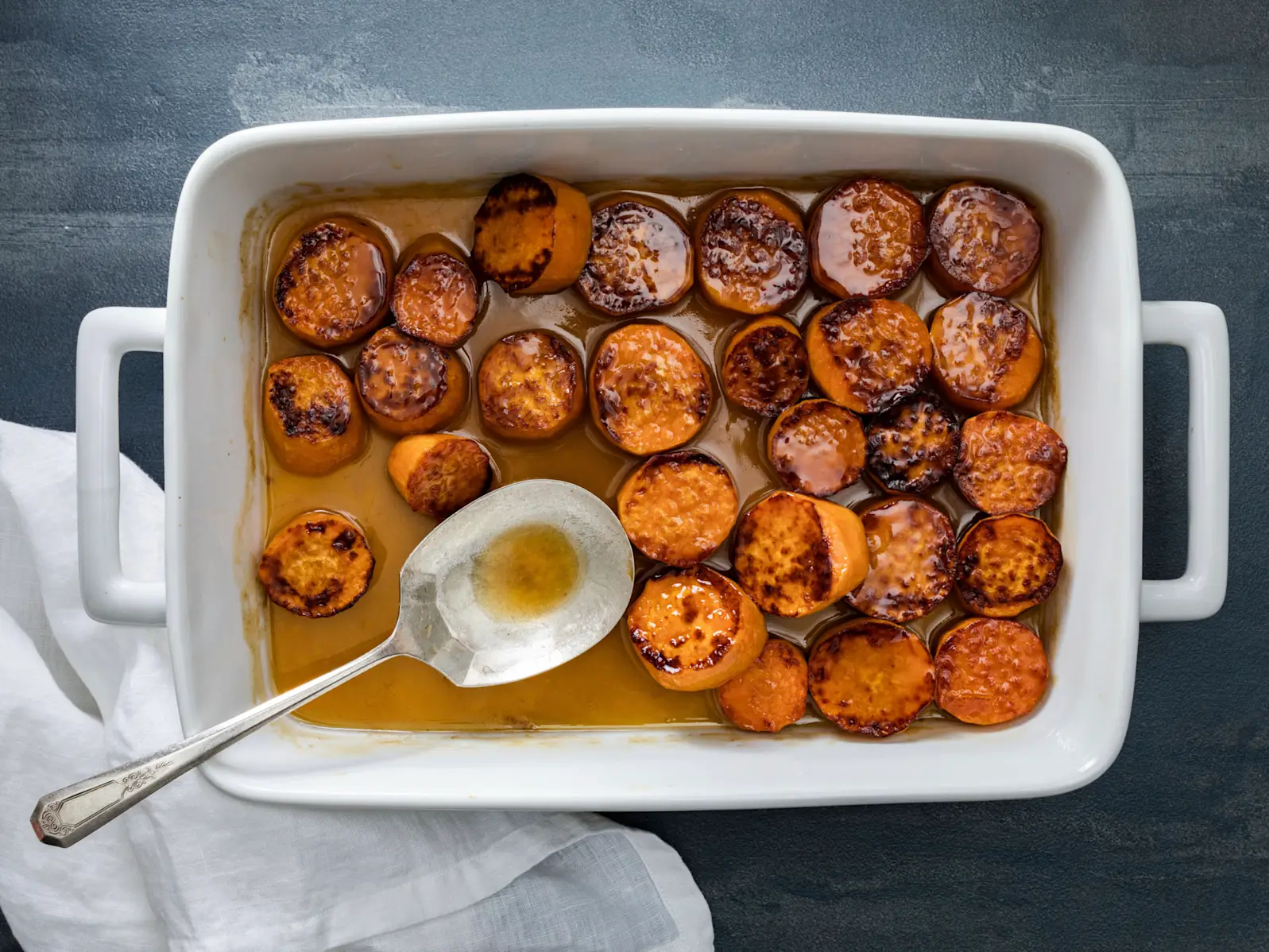 A white ceramic baking dish filled with glossy candied sweet potato chunks coated in amber-brown glaze, caramelized edges visible