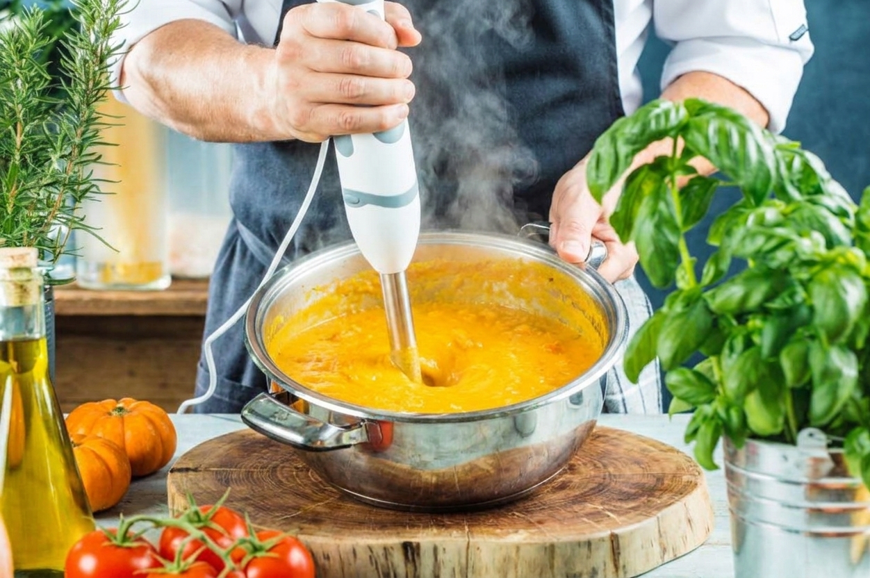 Hands holding an immersion blender in a pot of orange vegetable soup, with the surface turning smooth as it blends