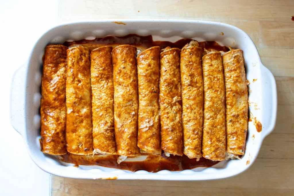 Hands rolling a filled corn tortilla into a tight enchilada over a baking dish with red sauce at the bottom