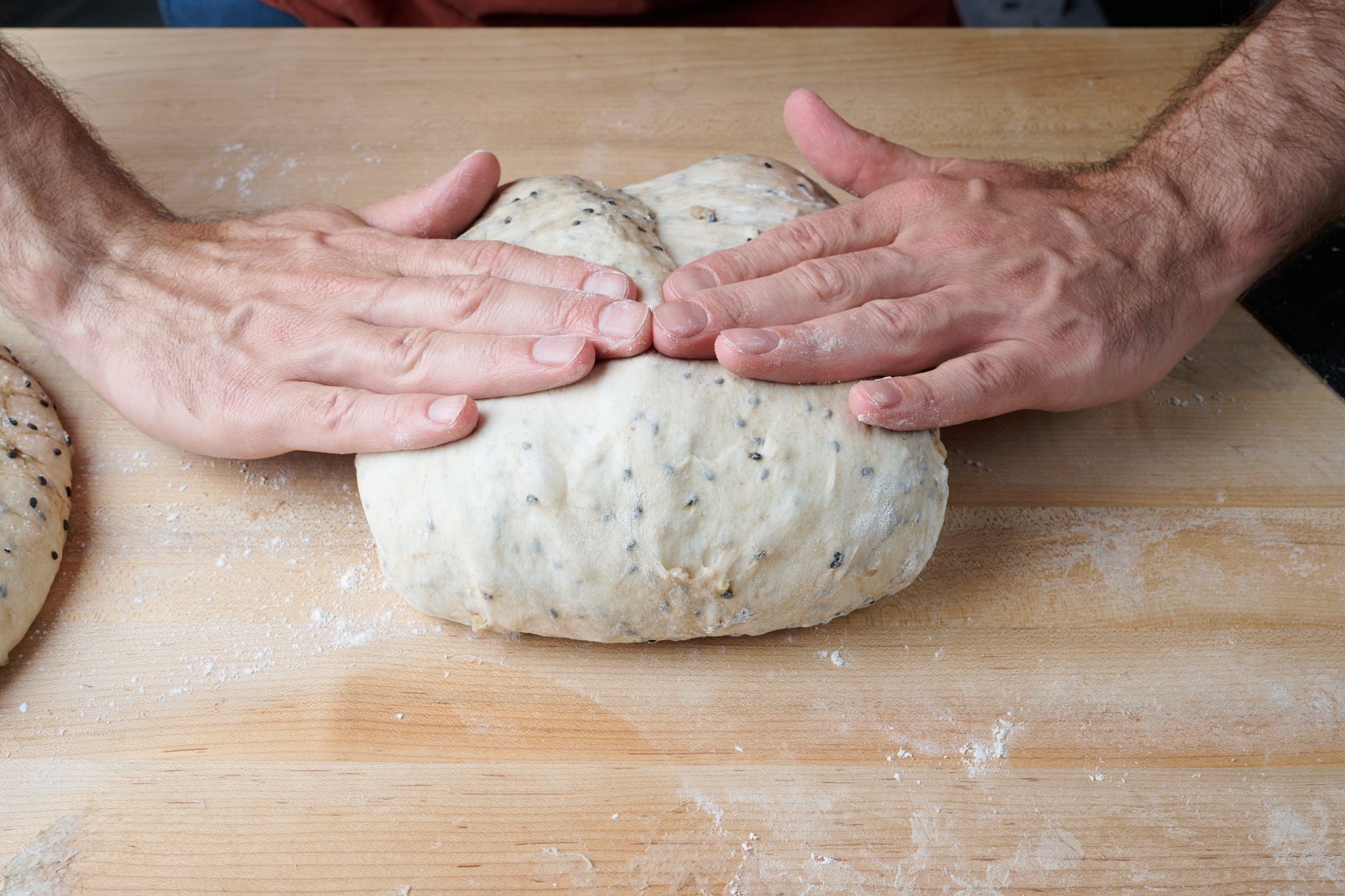 Hands rolling sourdough dough into a tight log on a lightly oiled countertop, preparing it for a loaf pan