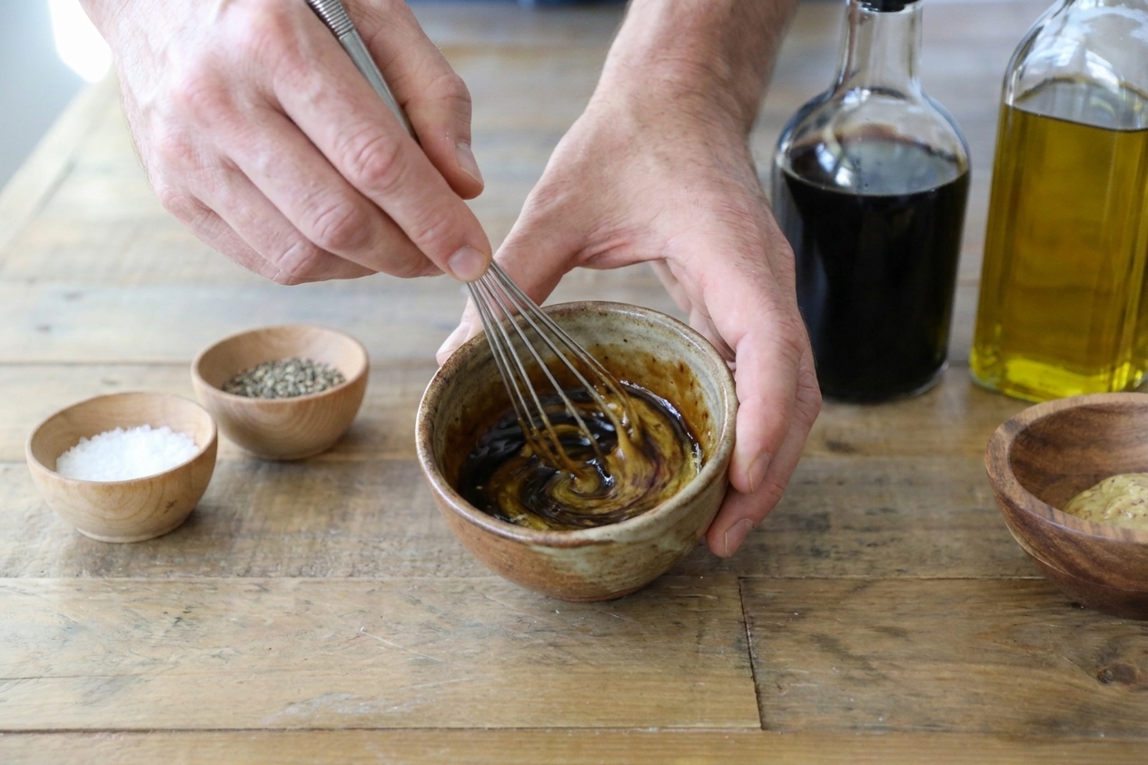 Hands whisking balsamic vinegar, Dijon, and olive oil together in a small ceramic bowl on a kitchen counter