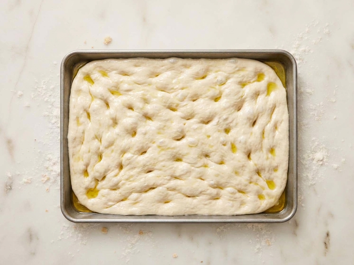 Pizza dough pressed into an oiled rectangular metal pan, with dimples from fingertips and oil glistening on the surface