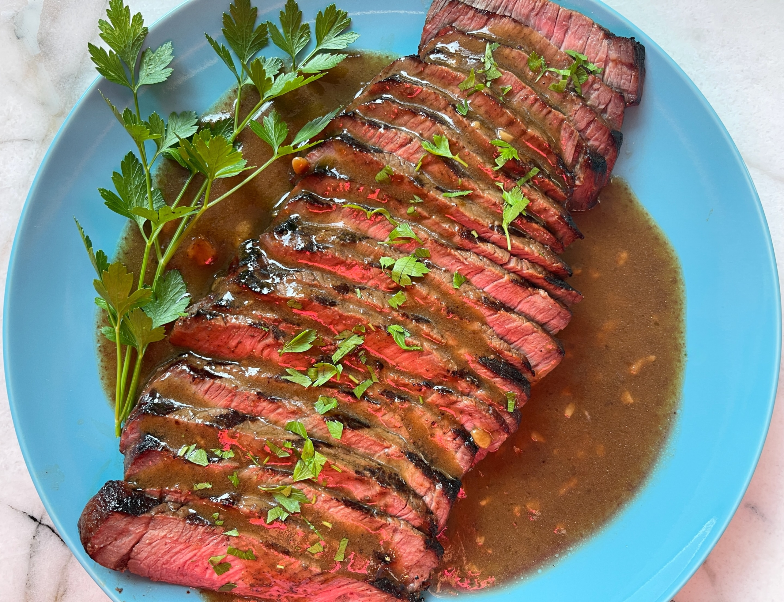 Thin slices of medium-rare London broil fanned on a platter with a glossy pan juice drizzle and chopped parsley