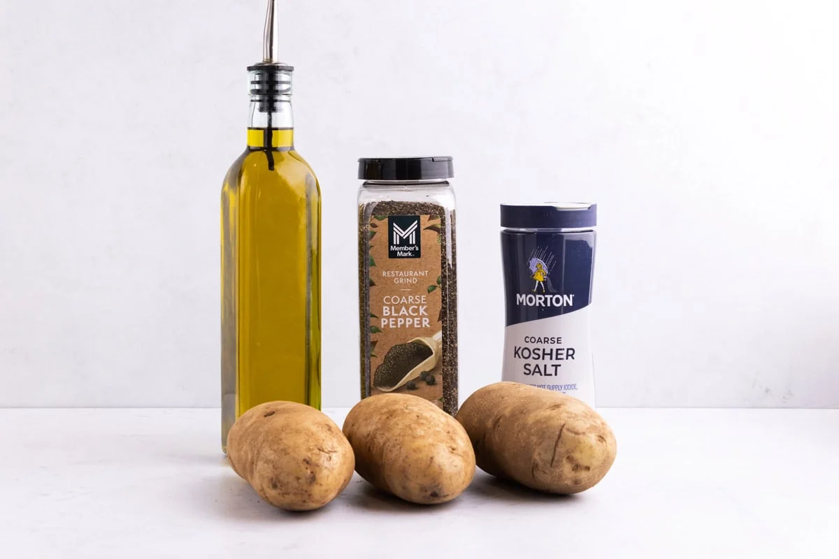 Whole russet potatoes on a kitchen counter next to a bowl of kosher salt and a small bottle of neutral oil